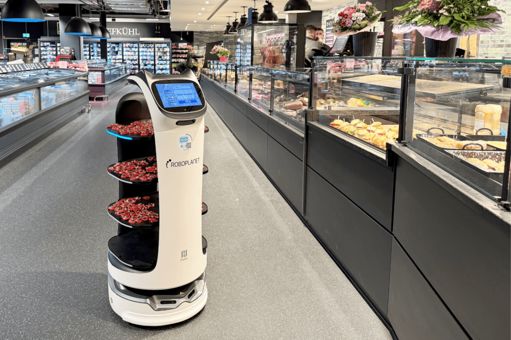 Pudu BellaBot von RoboPlanet mi Häppchen auf den Ablagen fährt durch Feinkost-Thekenbereich im Edeka